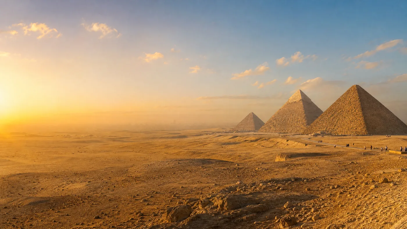 أهرامات الجيزة الثلاثة مصطفة في الساعة الذهبية من جهة الصحراء، في ضوء دافئ، مع بعض الزوار من بعيد